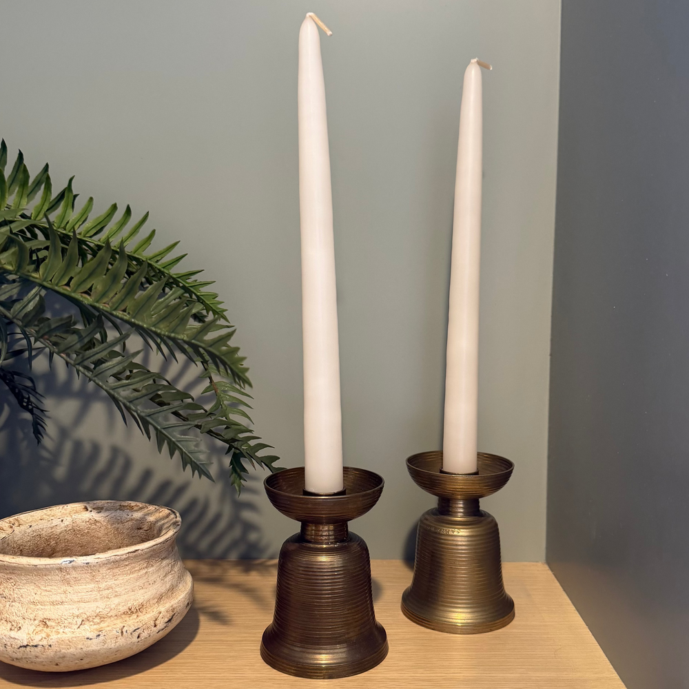 vintage brass candle holder with a tapered candle in it. green background on a wood shelf with a plant and bowl on the left side