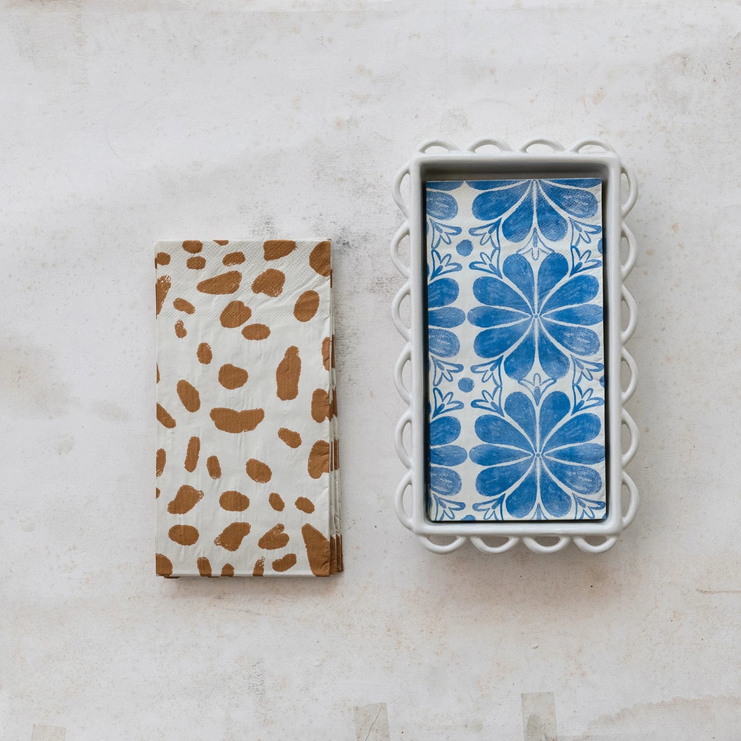Two patterned napkins on a marble surface, one with brown spots and the other with blue floral design in a white dish with scallop edges.
