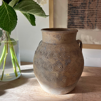 A rustic, earthy brown ceramic vase with embossed spiral patterns and side handles, displayed on a light wooden table next to a glass vase with white flowers.