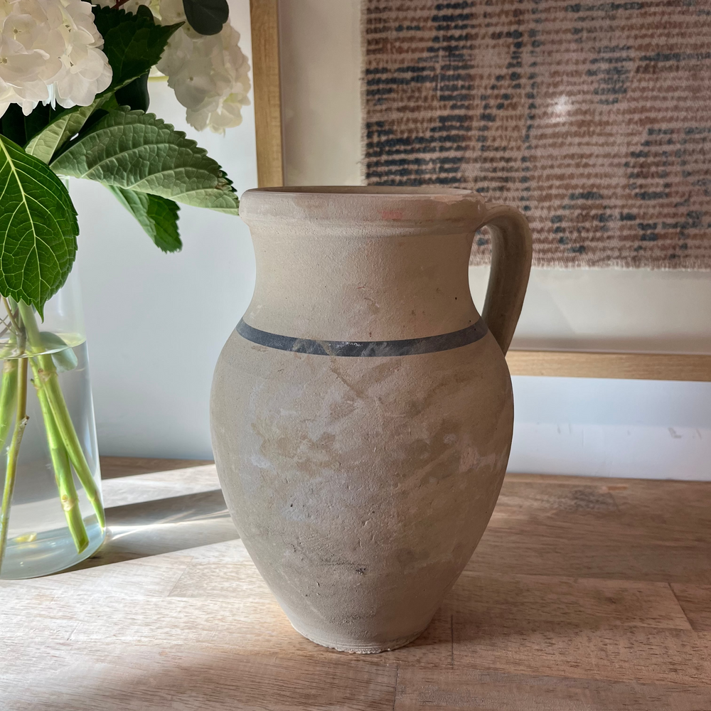 A rustic handmade ceramic vase with a handle, featuring a textured beige body and a dark gray band around the middle, sitting on a light wood tabletop next to a glass vase of white flowers.