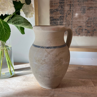A rustic handmade ceramic vase with a handle, featuring a textured beige body and a dark gray band around the middle, sitting on a light wood tabletop next to a glass vase of white flowers.