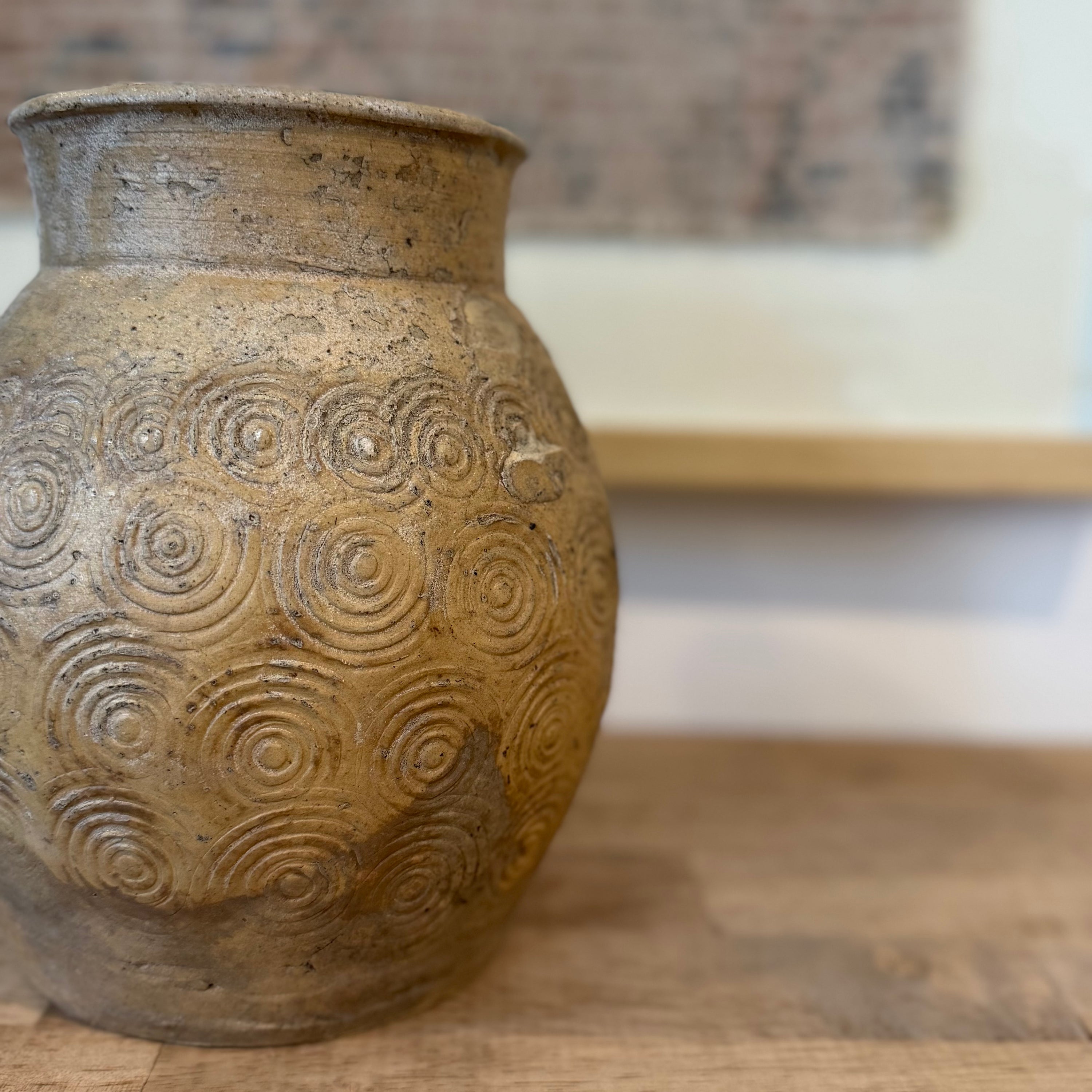 A handmade ceramic pot with a textured, earthy brown and beige finish, featuring a raised spiral pattern on the body, shown on a wooden surface with a blurred background.