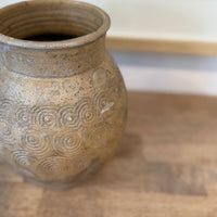 A close-up of a rustic ceramic vase with a textured, speckled beige and brown glaze, featuring embossed concentric spiral patterns on the front, set on a wooden surface.