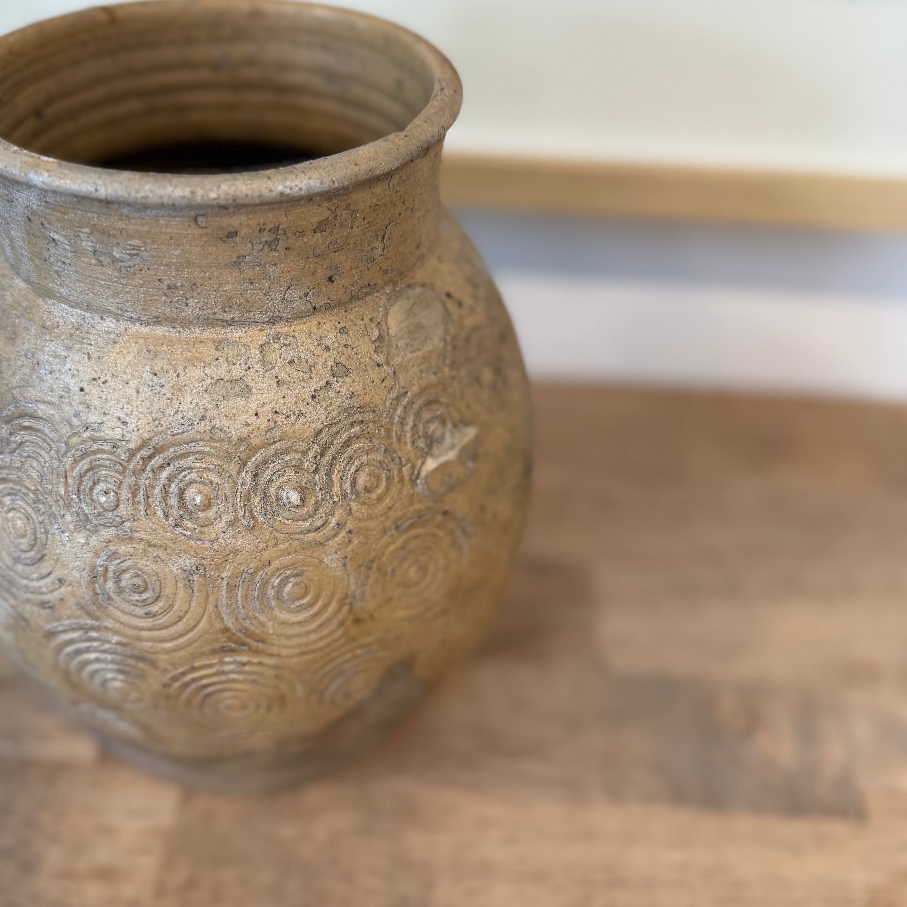 A close-up of a rustic ceramic vase with a textured, speckled beige and brown glaze, featuring embossed concentric spiral patterns on the front, set on a wooden surface.