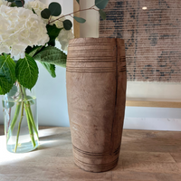 A tall, cylindrical wooden vase with carved horizontal grooves stands on a light wood floor next to a glass vase of white flowers and greenery, with a textured wall hanging in the background.