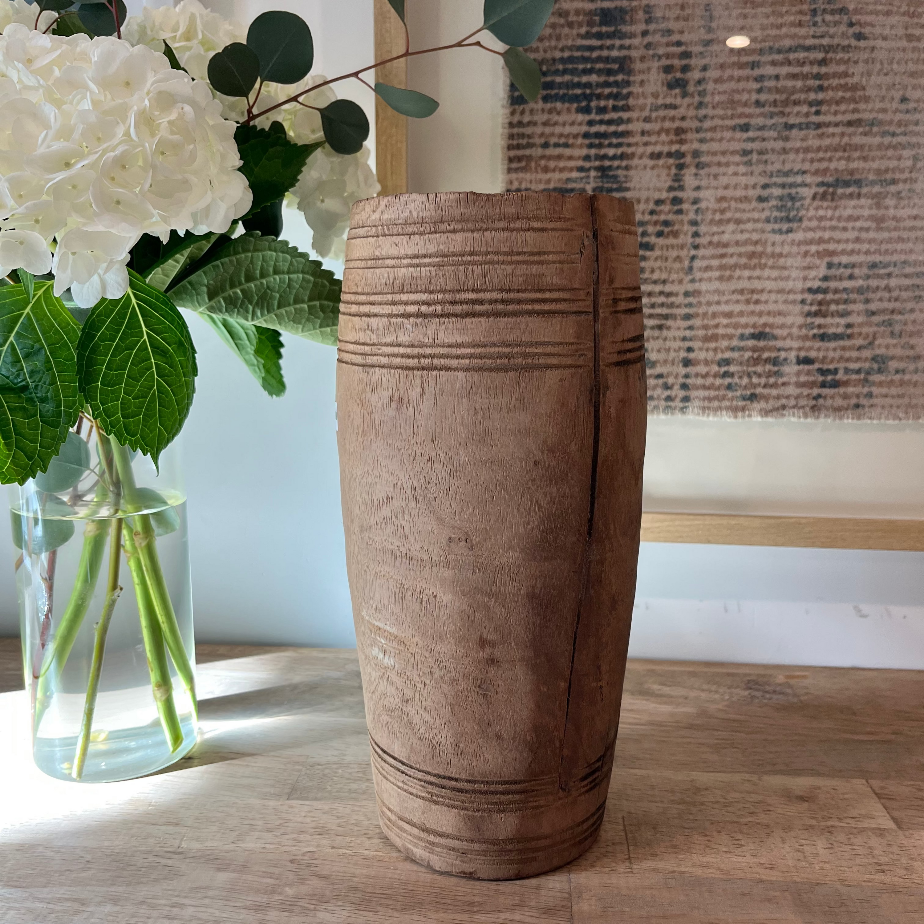 A tall, cylindrical wooden vase with carved horizontal grooves stands on a light wood floor next to a glass vase of white flowers and greenery, with a textured wall hanging in the background.