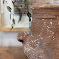 A close-up of a rustic terracotta clay pitcher with a small side spout, set on a wooden surface with a blurred background featuring a framed print and green leaves.