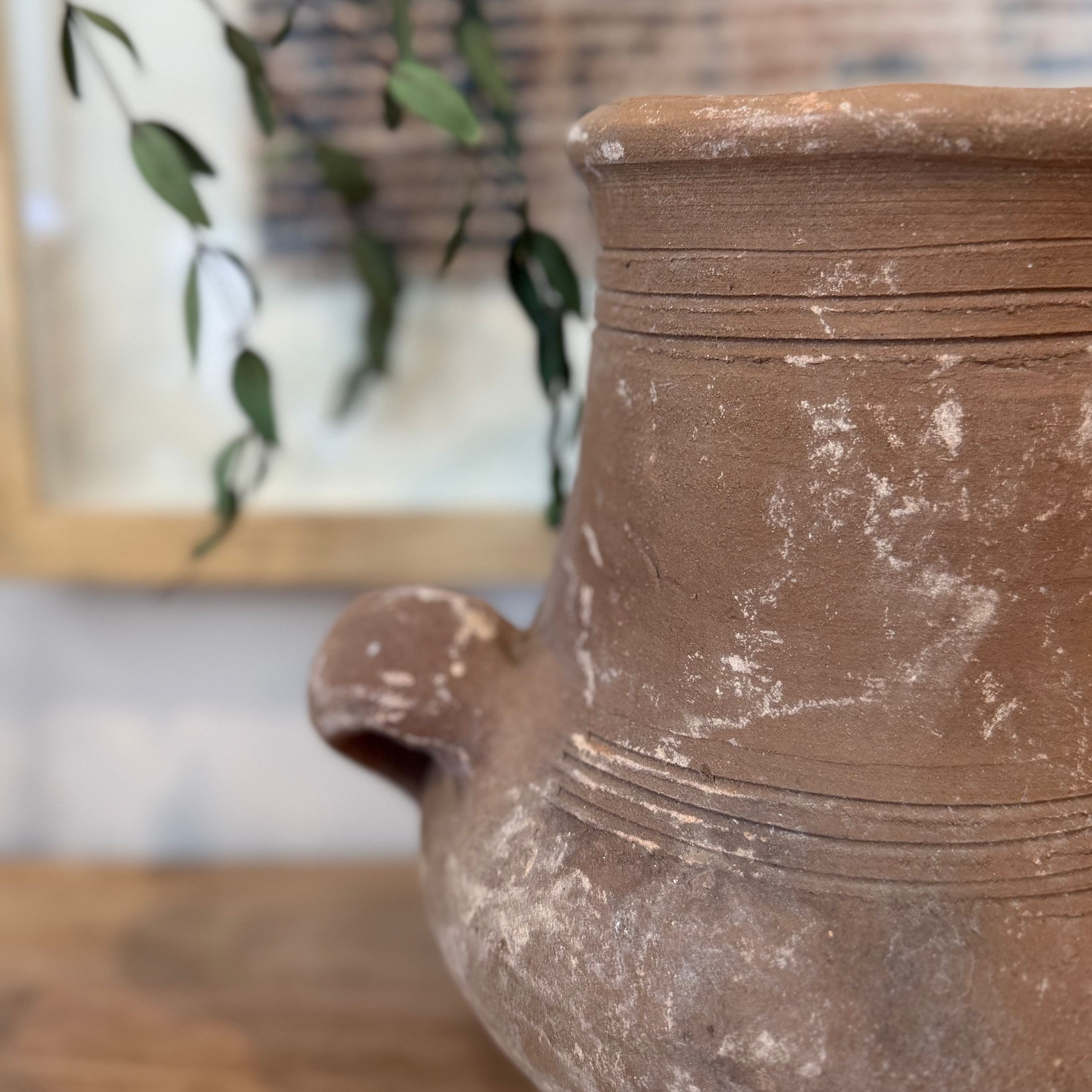 A close-up of a rustic terracotta clay pitcher with a small side spout, set on a wooden surface with a blurred background featuring a framed print and green leaves.