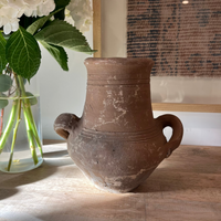 An antique-style brown terracotta vase with two side handles, displayed on a wooden table next to a glass vase of white flowers.
