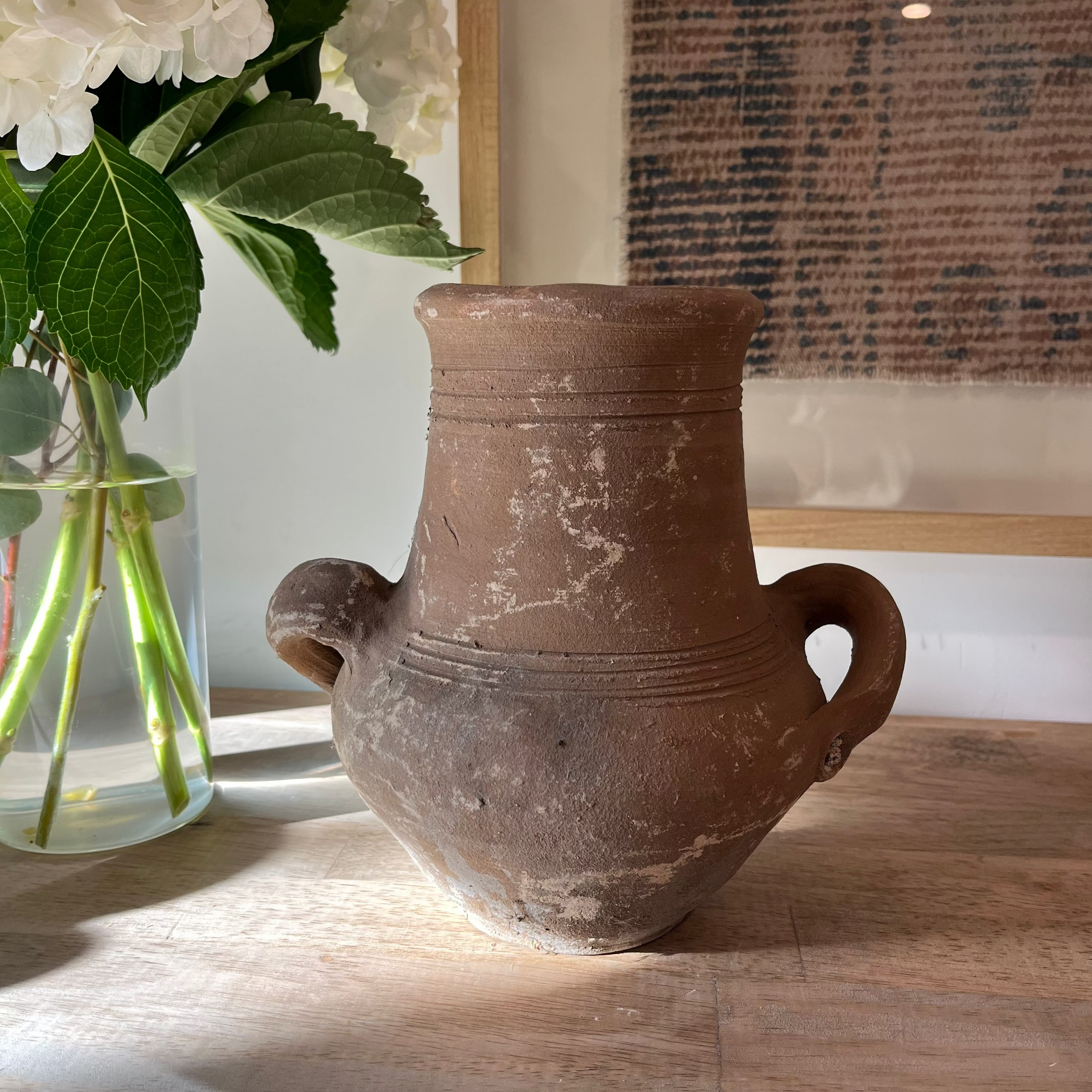 An antique-style brown terracotta vase with two side handles, displayed on a wooden table next to a glass vase of white flowers.