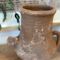 A rustic brown terracotta pot with a small  loop handle, resting on a wooden surface with a blurred indoor background.