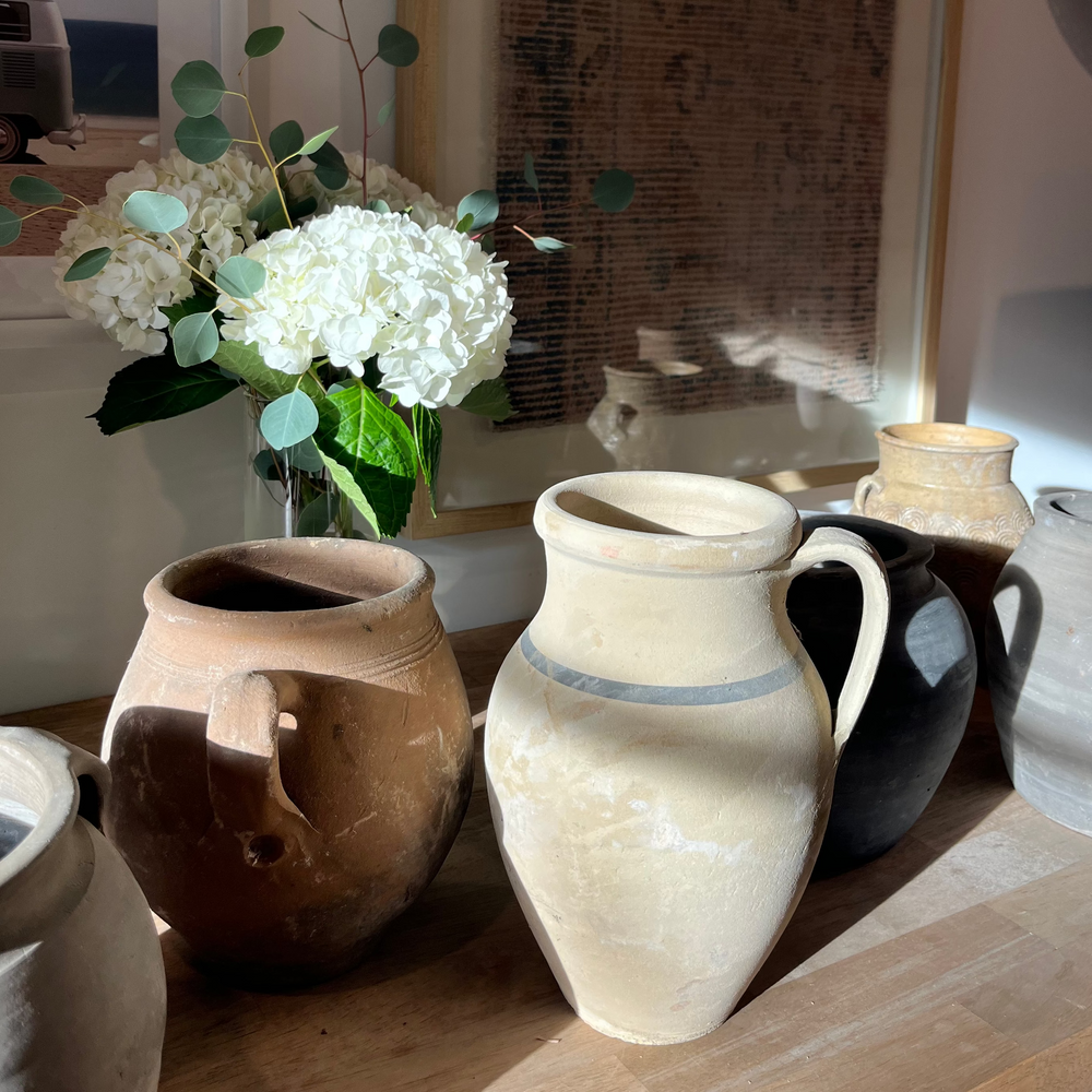 A collection of rustic handmade ceramic pitchers and vases in earthy tones of beige, brown, black, and gray, displayed on a wooden shelf next to a vase of white hydrangeas and eucalyptus.