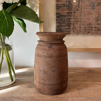 A rustic brown wooden vase with a flared rim and carved horizontal bands, displayed on a wooden tabletop next to a glass vase with white flowers.