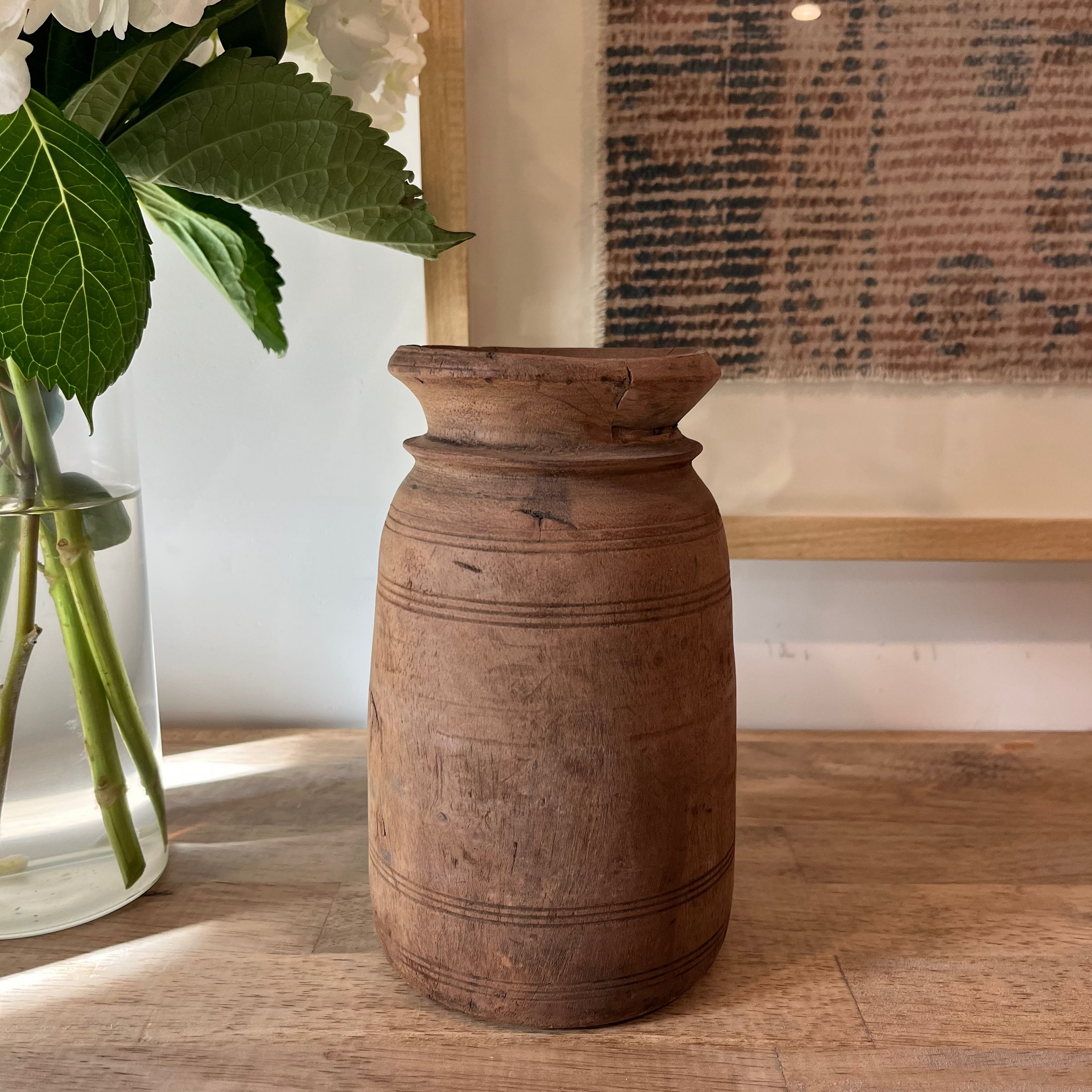 A rustic brown wooden vase with a flared rim and carved horizontal bands, displayed on a wooden tabletop next to a glass vase with white flowers.